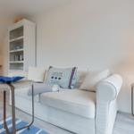 Weißes Sofa mit Kissen, Beistelltische und Bücherregal in einem hellen Raum.