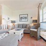 Wohnzimmer mit Sofa, Sessel und Fernseher. Terrakottafußboden und große Fenster.