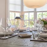 Gedeckter Esstisch mit Glas, Teller und Blumen vor Fenstern.