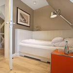 Schlafzimmer mit weißem Bett, orangefarbenem Nachttisch und Stehlampe unter schrägem Dach.