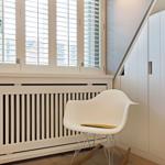 Weißer Stuhl mit Holzuntersitz vor Fenster mit Jalousien und Heizkörper.