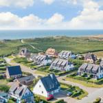 Houses with roofs and gardens near the coast. Sea and green hills in the background.