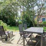 Terrace with table and chairs, surrounded by greenery and a house in the background.