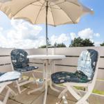 Terrasse mit Tisch, drei Stühlen und Sonnenschirm. Weißer Holzzaun und Bäume im Hintergrund.