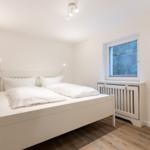 Ein Schlafzimmer mit weißem Bett, Holzboden und Fenster mit Heizkörper.