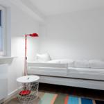 Ein Schlafzimmer mit weißem Bett, roter Stehlampe und buntem Teppich.