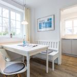 Essbereich mit weißem Tisch, Stuhl und Bank. Küche im Hintergrund. Holzboden und Fenster.