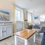 Wohnzimmer mit Holztisch, grauen Sesseln und weißen Schränken. Fenster mit Vorhängen.