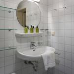 Badezimmer mit Waschbecken, Spiegel und Glasregalen an weißen Fliesenwänden.