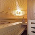 Sauna mit Holzbank und Eimer. Warmes Licht. Wanduhr und Heizung sichtbar.