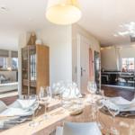 Esszimmer mit Tisch, Stühlen und Gläsern. Küche und Wohnzimmer im Hintergrund.