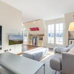 Wohnzimmer mit Sofa, Couchtisch und Fernseher. Durchgang zur Küche und Terrasse.