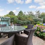 Garden with wicker furniture, glass table, and green-and-white lounge chair. Flower beds and trees in the background.
