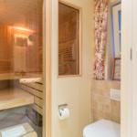 Sauna with wooden bench and glass wall, next to toilet and window with curtain.