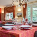 Dining table with tablecloth, glasses, and cutlery. Windows with curtains and chandelier.
