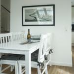Dining room with white table and chairs, wall picture, and kitchen area in background.