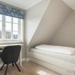 Ein Schlafzimmer mit Dachschräge, weißem Bett und Schreibtisch unter dem Fenster.