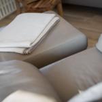 Graue Ledercouch mit gefaltetem Deckenbezug und Holzboden im Hintergrund.