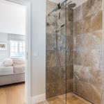 Shower with glass enclosure and stone tiles. Bedroom with bed and window visible in background.