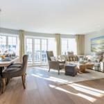Living and dining area with wooden floor, sofa, table and chairs, large windows.