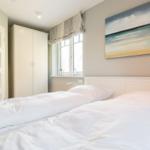 Schlafzimmer mit weißem Bett, Kleiderschrank und Fenster. Wandbild zeigt Meereslandschaft.