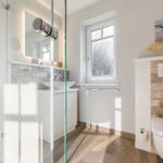 Badezimmer mit Dusche, Waschbecken und WC. Holzboden und Fenster.