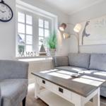 Wohnzimmer mit grauem Sofa, Tisch und Wanduhr. Fenster mit Pflanze und Wandbild.
