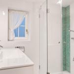 Badezimmer mit Dusche, Waschbecken und Fenster mit weißen Vorhängen.