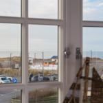Durch das Fenster blickt man auf einen Strandparkplatz und das Meer.
