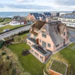 Haus mit strohgedecktem Dach und Glasbrüstung, umgeben von Grünflächen und Strand.