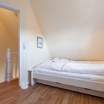 Schlafzimmer mit weißem Bett, Holzboden und schrägen Wänden. Tür führt zu Treppenhaus.