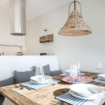 Dining area with rustic wooden table and gray bench. Hanging lamp and kitchen area in background.
