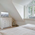 Schlafzimmer mit Dachschrägen, weißem Bett und TV-Schrank. Fenster mit Blick auf Bäume.