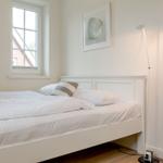 Ein Bett mit weißer Bettwäsche, Wandbild und Stehlampe neben einem Fenster.