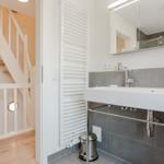 Badezimmer mit Waschbecken, Spiegel und Heizkörper. Treppenhaus im Hintergrund.