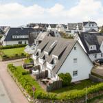White house with gray roof and garden. Street with cars and view of the sea.