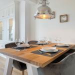 Dining room with wooden table, chairs, and pendant light. Kitchen cabinets in background.