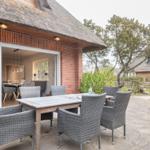 Terrasse mit Tisch und Stühlen vor einem Haus mit Strohdach und Glasfront.