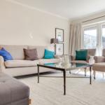 Living room with sofa, two armchairs, and glass table. Windows with curtains. Rug and lighting.