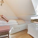 Schlafzimmer mit Dachschrägen, Bett, Wickerstuhl und Fernseher auf weißen Möbeln.