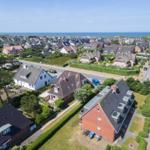 Aerial view of a coastal residential area with houses, greenery, and the sea in the background.