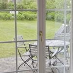 Through the glass door, a paved balcony with table and chairs is visible. Behind it lies a green lawn.