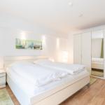 Schlafzimmer mit weißem Bett, Nachttisch und Kleiderschrank. Holzboden und Wandbild.
