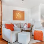 Living room with white sofa, orange cushions and ottomans. Wall art and plants complete the space.
