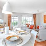 Dining room with round table, white chairs, and orange curtains. Living area with sofa and wall art.