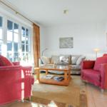 Wohnzimmer mit roten Sesseln, Sofa und Holztisch. Fenster mit Vorhängen.
