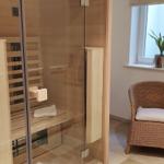 Sauna mit Glaswand und Sitzbank. Nebenher steht ein Rattan-Stuhl mit Kissen.