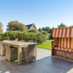Terrasse mit Esstisch, Stühlen und Strandkorb. Grünflächen und Häuser im Hintergrund.