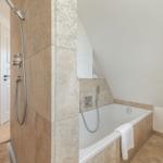 Bath with shower and bathtub, tiled with stone and wooden stool.