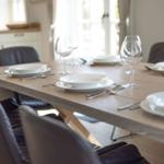 Wooden table with white plates and glasses, dark chairs, kitchen in background